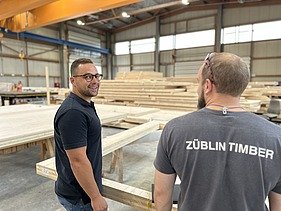 Foto von zwei jungen Männern, die in der Produktion eines Holzbaubetriebs stehen.