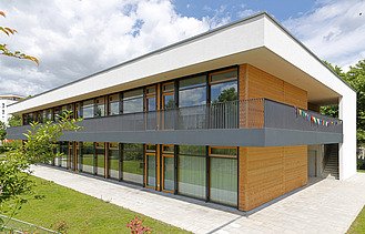 Foto einer Außenaufnahme der Kindertagesstätte in Frankfurt. Das Holzgebäude hat ein Flachdach und eine Holz- Glasfassade.