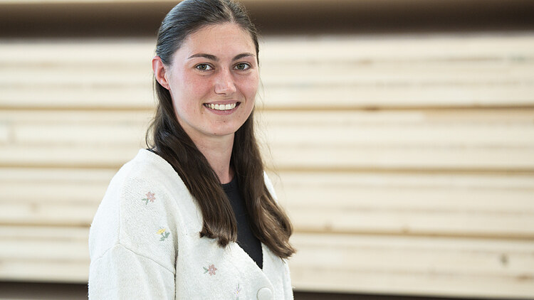 Technical office at ZÜBLIN Timber Picture of a young woman with dark long hair, smiling and wearing a white cardigan. In the background is a pile of wood