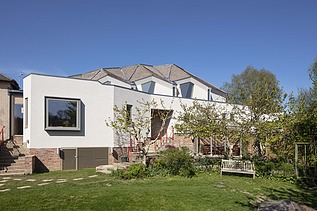 Foto eines Gebäudes mit weißer Fassade und Dach mit Gauben. Blauer Himmel und Bäume sind zu sehen.