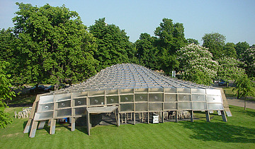 Foto des Serpentine Pavillons in London. Man sieht eine offene, begehbare Holzkonstruktion auf einer grünen Wiese mit einem Baum.