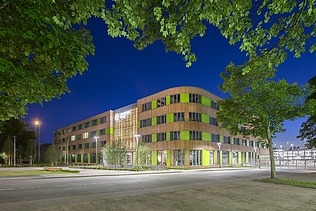 Foto einer Außenaufnahme bei Nacht. Man sieht ein beleuchtetes großes Holzgebäude mit Holzfassade und grünen Fassadenelementen.