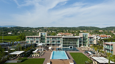 Photo of the Hotel Aqualux ini Bardolino. You can see a large building with a glass façade and flat roof, as well as a pool.