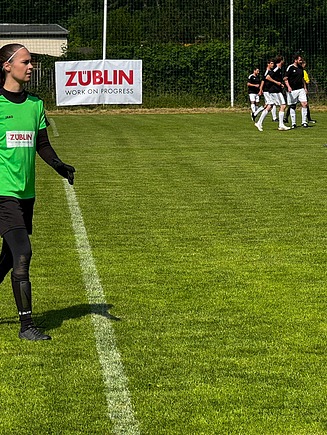 Foto von einem Fußballplatz mit Sportlern darauf. Eine junge Frau in grünem Towarttrikot geht über das Feld.