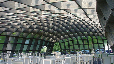 Foto des bestuhlten Innenraumes des Serpentine Pavillons in London. Man sieht eine schöne Holzkonstruktion des Daches.