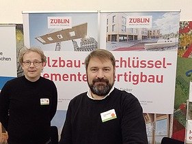 Foto von zwei Männern. Im Hintergrund sind Ausschnitte zweier Roll-Ups zu sehen. 