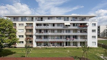 Photo of a multi-storey building with garden and hard court in front. Wooden extension.
