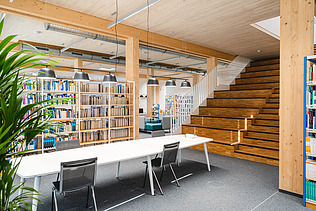 Foto der Bibliothek der Uni Witten. Man sieht viele Bücher, eine Holzdecke und Holztreppe.