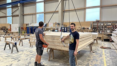 Foto von zwei Männern in der Produktionshalle. Sie stehen vor einem Stapel voller Holzelemente, welche am Kran hängen.