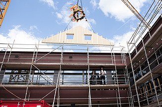 Photo of a wooden gable wall element suspended from a crane above the Schule am Hangs in Frankfurt.