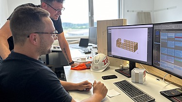 Photo of two men at a computer workstation. One is sitting, the other is standing.