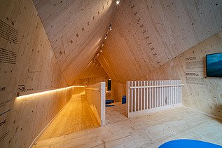 Foto des Innenraums des Holzpavillons NEXT500 der Fuggerei Augsburg. Man sieht einen Raum mit vielen Holzoberflächen.
