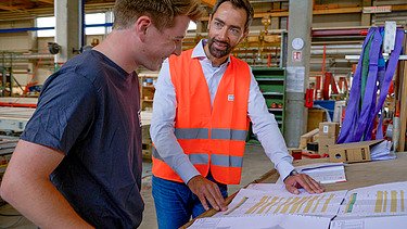 Foto von zwei jungen Männern in der Produktionshalle von ZÜBLIN Timber. Sie stehen vor einem Bauplan und reden miteinander.