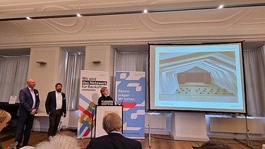 Foto von drei Personen, die ein Projekt präsentieren und vor Publikum stehen.