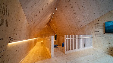 Foto des Innenraums des Holzpavillons NEXT500 der Fuggerei Augsburg. Man sieht einen Raum mit vielen Holzoberflächen.