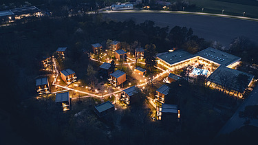 Photo of an aerial view at night. You can see illuminated houses and trees.