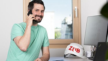 Foto eines Mannes am Bildschirmarbeitsplatz mit dem Handy am Ohr.