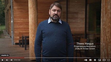 Nahaufnahme eines Mannes, der in die Kamera blickt. Im Hintergrund ein Holzgebäude.