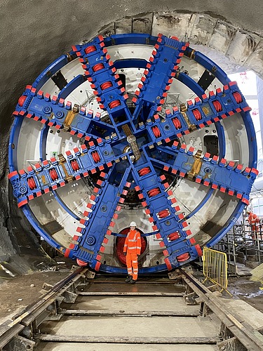 Foto von einer jungen Frau in oranger Arbeitskleidung vor einer rießengroßen Tunnelbohrmaschine in blau-rot.