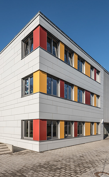 Außenaufnahme der Schule in Erfurt. Man sieht ein großes, weißes Gebäude mit bunten Farbelementen bei den Fenstern.