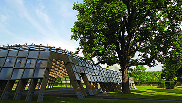 Foto des Serpentine Pavillons in London. Man sieht eine offene, begehbare Holzkonstruktion auf einer grünen Wiese mit einem Baum.