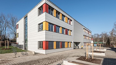 Außenaufnahme der Schule in Erfurt. Man sieht ein großes, weißes Gebäude mit bunten Farbelementen bei den Fenstern.