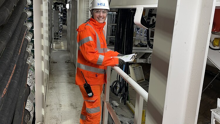 Trainee and site manager at ZÜBLIN Timber Picture of a young woman in construction site clothing with a white helmet on her head. She is standing on a railing in a building.