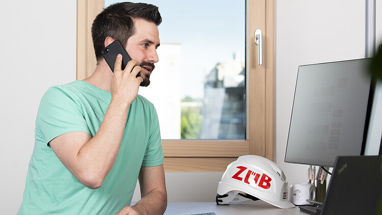 Work preparation at ZÜBLIN Timber Photo of a young man with dark hair and a short beard standing at a PC workstation.