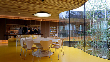 Foto eines Innenraumes mit großer Glasfensterfront, Holzdecke und Tisch mit Stühlen.