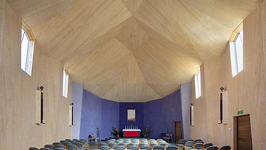 Foto eines Innenraums einer Kapelle mit großer gewölbter Holzdecke. Eine Seite hat eine blaue Wand.