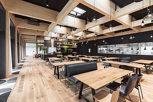 Foto eines Aufenthaltraumes mit Holztischen und Stühlen. Holzboden und Holzdecke.