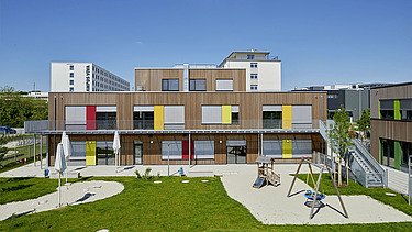 Foto eines Kindergartens in München. Zu sehen ist ein Gebäude aus Holz mit Garten.