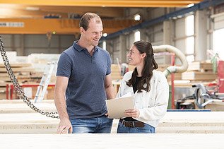 Foto eines großen Mannes und einer jungen Frau mit dunklen, langen Haaren, die in einer Produktionshalle stehen und sich anschauen.