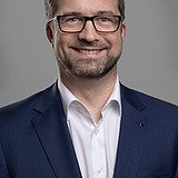 Foto eines lachenden Mannes mit Vollbart und Brille, blauem Jackett und weißem Hemd. Rainer Stettberger.