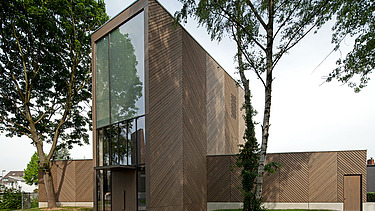 Foto einer Außenaufnahme der Immanuelkirche in Köln. Diese hat eine Holzfassade und ein Flachdach und einen je seitlichen Anbau aus Holz.
