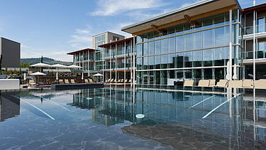 Photo of the Hotel Aqualux ini Bardolino. You can see a large building with a glass façade and flat roof, as well as a pool.