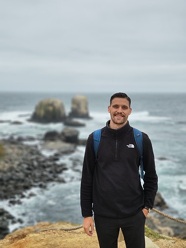 The photo shows a young man in black clothing. He is standing in the sand. In the background you can see the sea and rocks.