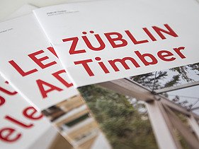 Foto von drei Coverseiten der Broschüren von ZÜBLIN Timber. Man sieht große, rote Aufschriften 
