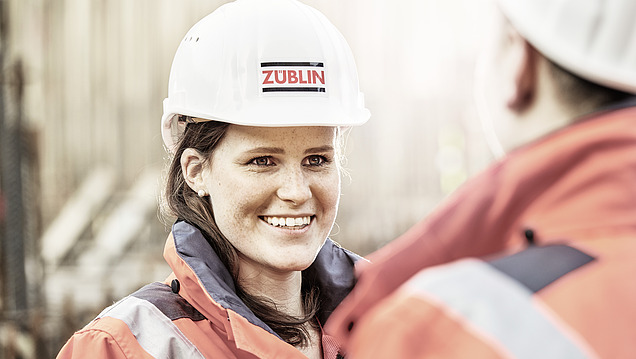 Photo of a young woman with a white construction helmet on and in work clothes. She is talking to a man.