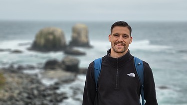 The photo shows a young man in black clothing. He is standing in the sand. In the background you can see the sea and rocks.