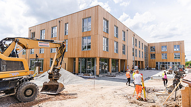 Photo of Witten University from the outside. You can see a large wooden building, an excavator and people.