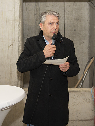 Foto von Simon Pfeffer, der ins Mikrofon Grußworte spricht.
