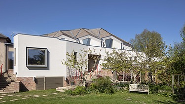 Foto eines Gebäudes mit weißer Fassade und Dach mit Gauben. Blauer Himmel und Bäume sind zu sehen.