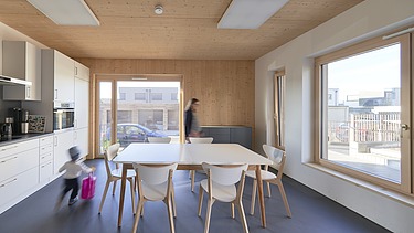 Innenaufnahme eines möblierten Raumes einer Küche. Man sieht viele Holzoberflächen und große Fensterflächen.