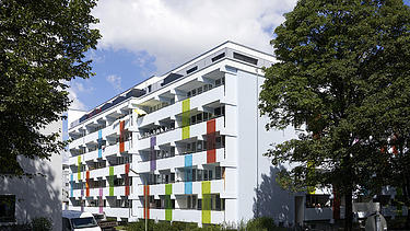Photo of a multi-storey building with a wooden extension. The façade is white with colorful façade elements.
