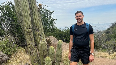 Photo of a young man in short black trousers and a black T-shirt and he is standing in the desert. He is standing next to a large cactus.