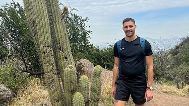 Foto eines jungen Mannes in kurzer schwarzer Hose und schwarzem T-Shirt und er steht in der Wüste. Er steht neben einem großen Kaktus.