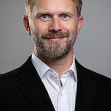 Kontakt ZÜBLIN Timber: Tilman Conrad Foto eines lachenden Mannes mit Vollbart, schwarzem Jackett und weißem Hemd. Tilmann Conrad.