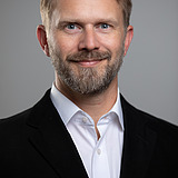 Contact ZÜBLIN Timber: Tilman Conrad Photo of a smiling man with a full beard, black jacket and white shirt. Tilmann Conrad.
