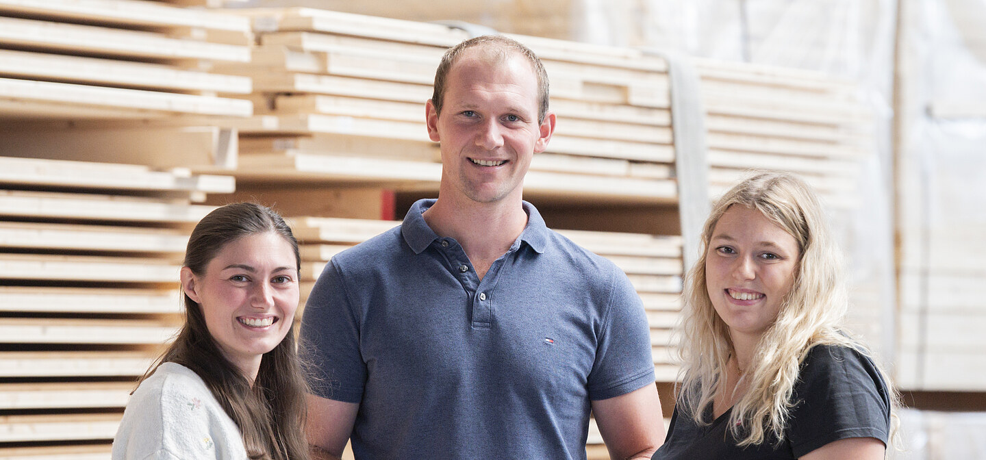 Karriere ZÜBLIN Timber Foto von drei jungen Personen, die vor einem LENO-Brettsperrholz-Stapel stehen.